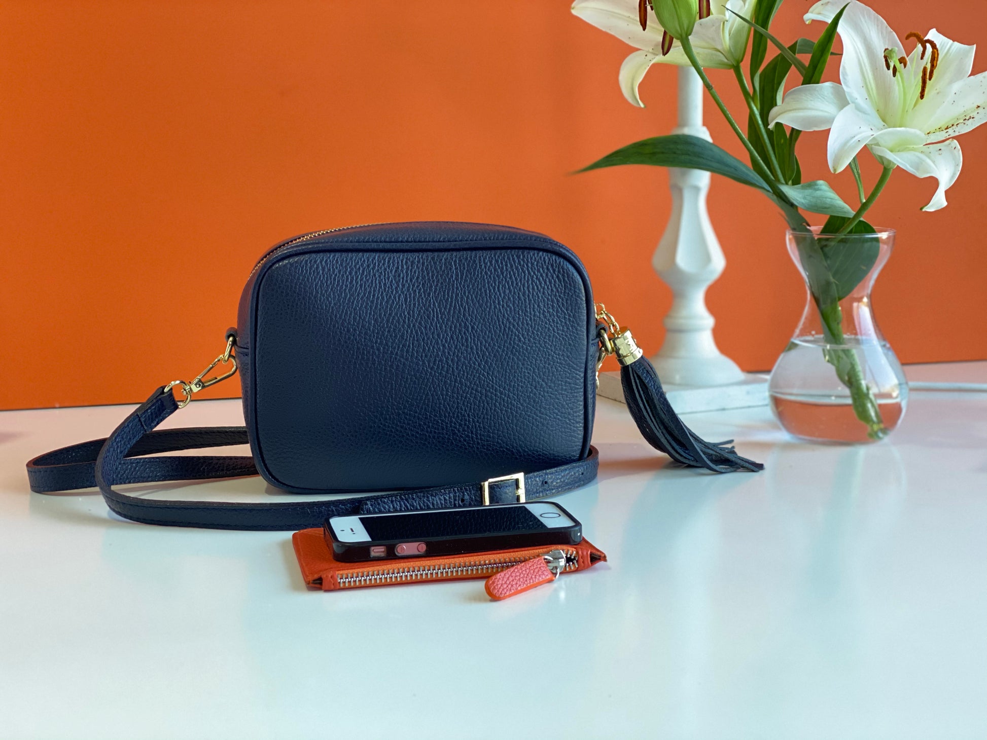 An image of a navy blue leather messenger bag with an orange purse and a mobile phone in front of it. There is a  vase of lilies next to the bag.